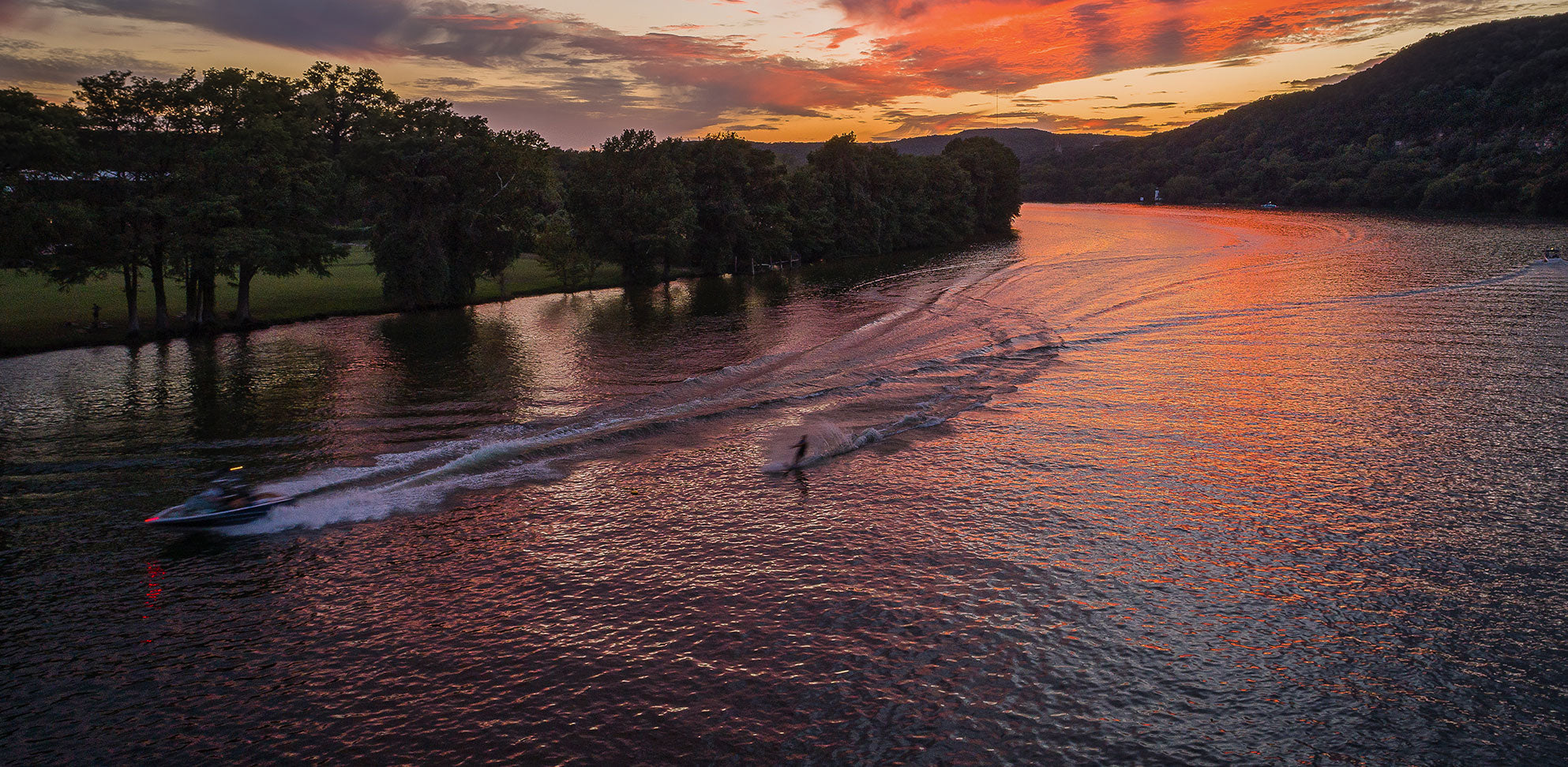 Boat towing a waterskier at sunset, leaving a long wake as the sky reflects in the water.