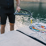 Person in a wetsuit standing on a dock, holding the black handle with a coiled multicolored rope beside them.