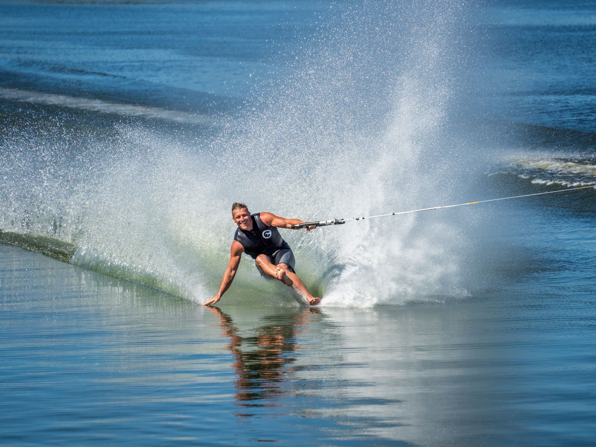 Barefoot waterskier carving across glassy water, creating a large spray while gripping the tow rope.
