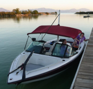 White and maroon boat docked at a pier with a red bimini top, occupied by two people near a calm lake.