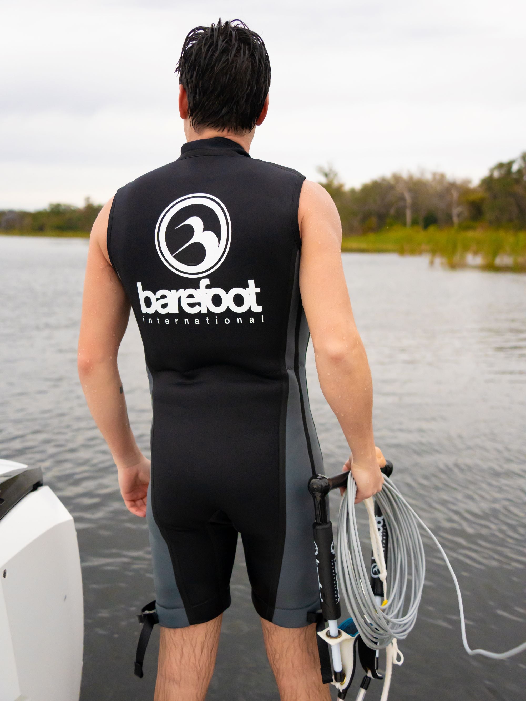 A man standing near the water wearing the Iron Sleeveless Wetsuit, holding a boom handle and coiled cable.