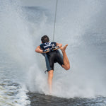 A barefoot waterskier wearing the Iron Sleeveless Wetsuit, balancing on one foot while creating a large water spray.