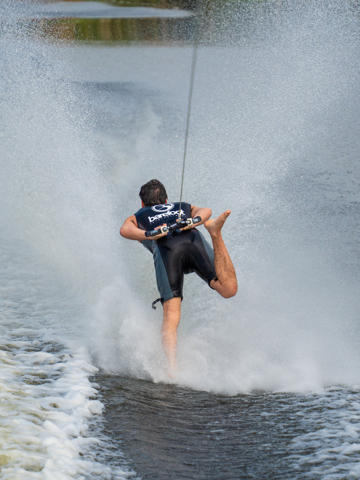 A barefoot waterskier wearing the Iron Sleeveless Wetsuit, balancing on one foot while creating a large water spray.