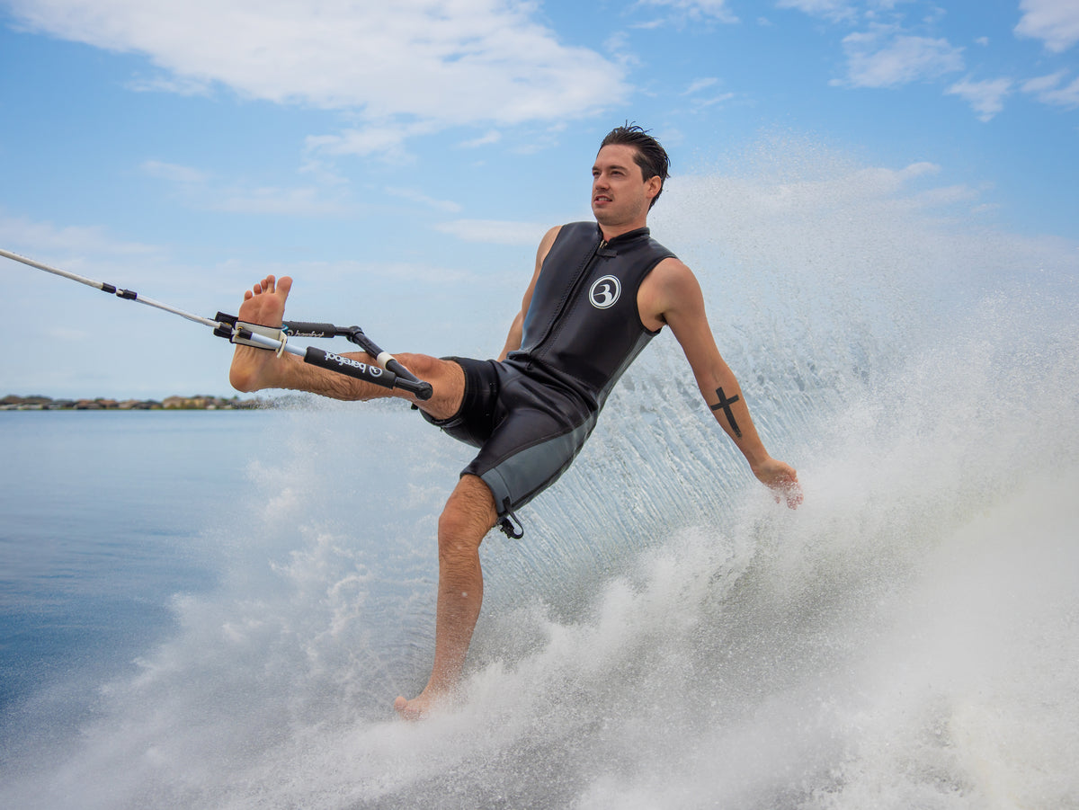 A barefoot waterskier wearing the Iron Sleeveless wetsuit performs a trick on the water, kicking up a spray.