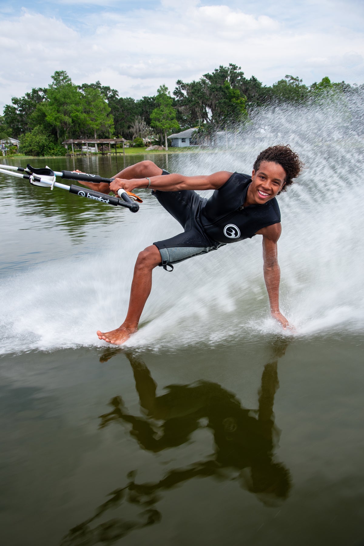 Person barefoot waterskiing on one foot, one hand dragging in the water, smiling on a lake with trees and houses in the background