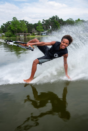 Person barefoot waterskiing on one foot, one hand dragging in the water, smiling on a lake with trees and houses in the background
