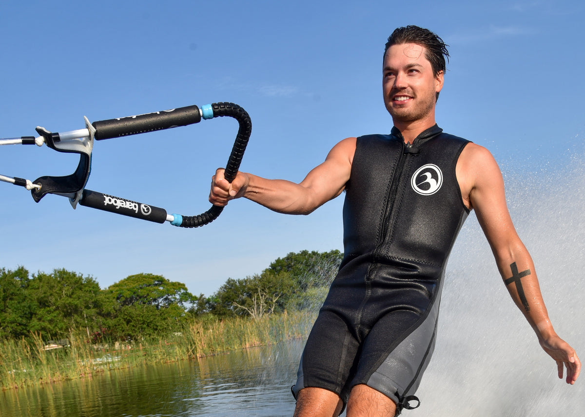 Man barefoot water skiing on a lake with trees in the background wearing a barefoot international wetsuit and holding a barefoot international rope and handle