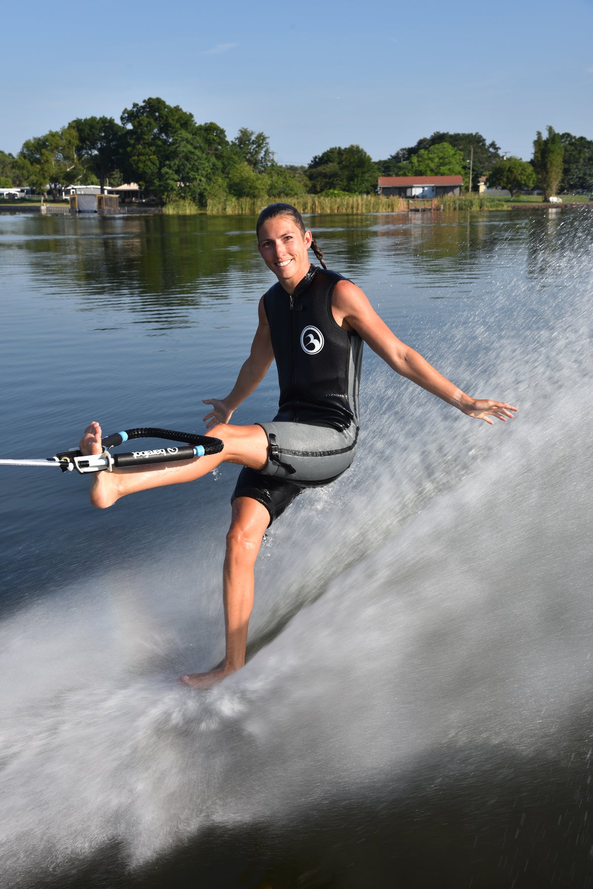 Person barefoot waterskiing on one foot, holding the rope with his foot, smiling on a lake with trees and houses in the background