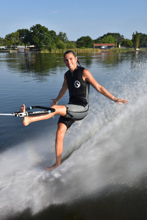 Person barefoot waterskiing on one foot, holding the rope with his foot, smiling on a lake with trees and houses in the background