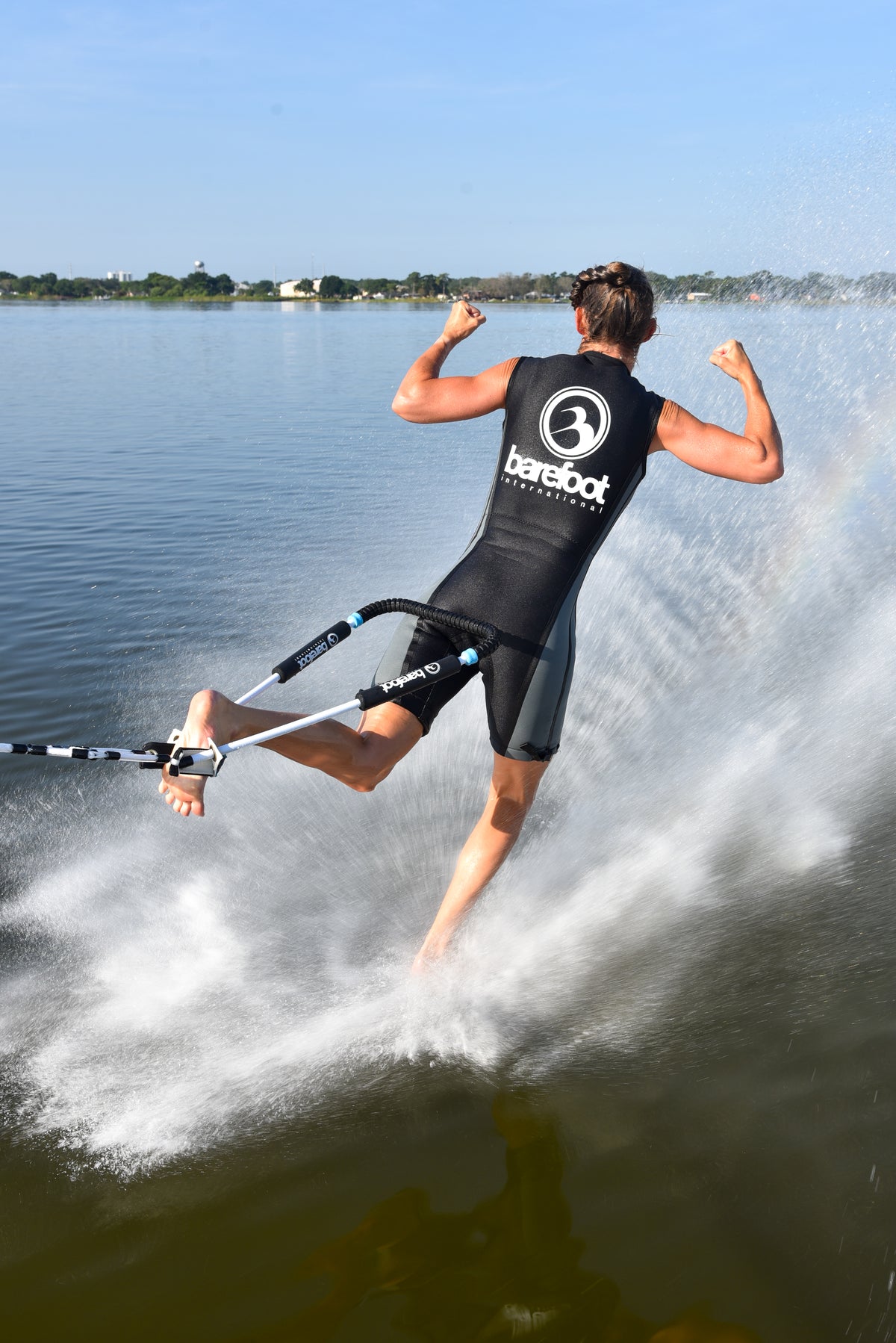 Person barefoot water skiing on a lake standing on one foot, flexing their ars wearing a life vest with 'barefoot' branding