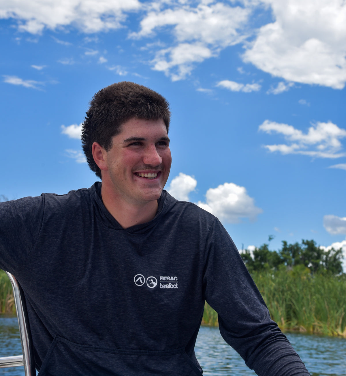 Smiling man wearing a gray long-sleeve UV protection sun shirt on a boat by a lake.