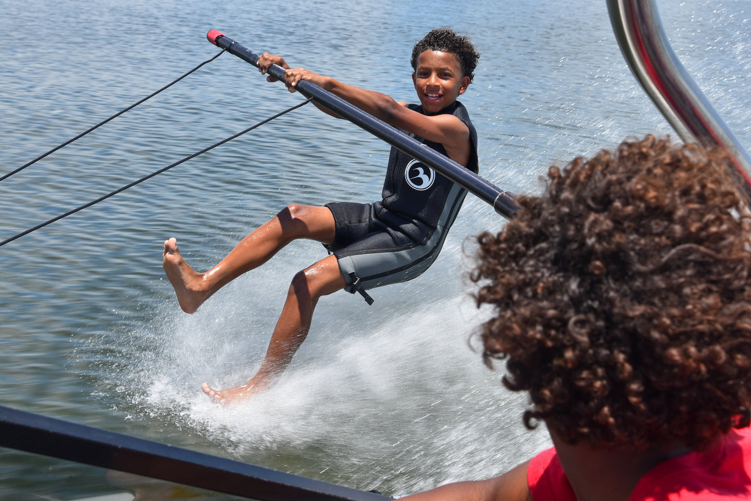 Smiling boy barefoot skiing on a boom in the Juniors black and gray Iron Sleeveless Wetsuit.