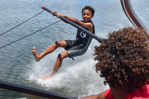 Smiling boy barefoot skiing on a boom in the Juniors black and gray Iron Sleeveless Wetsuit.