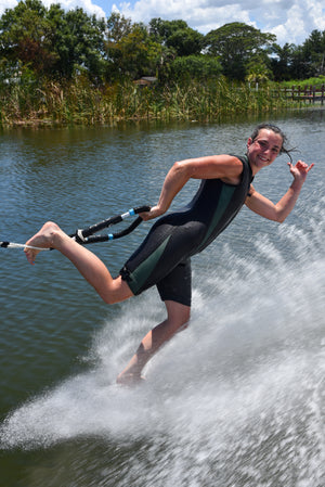 Woman barefoot skiing in a black and green Iron Sleeveless Wetsuit, spraying water.