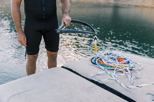 Person in a wetsuit standing on a dock, holding the black handle with a coiled multicolored rope beside them.